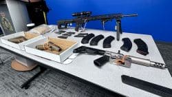 Table spread with multiple firearms and magazines laid out for display or evidence collection in a room with a blue wall behind.