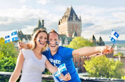 Couple smiling and waving small Quebec flags on a balcony with a château-like hotel in the background; face painted in blue fleur-de-lis symbols.
