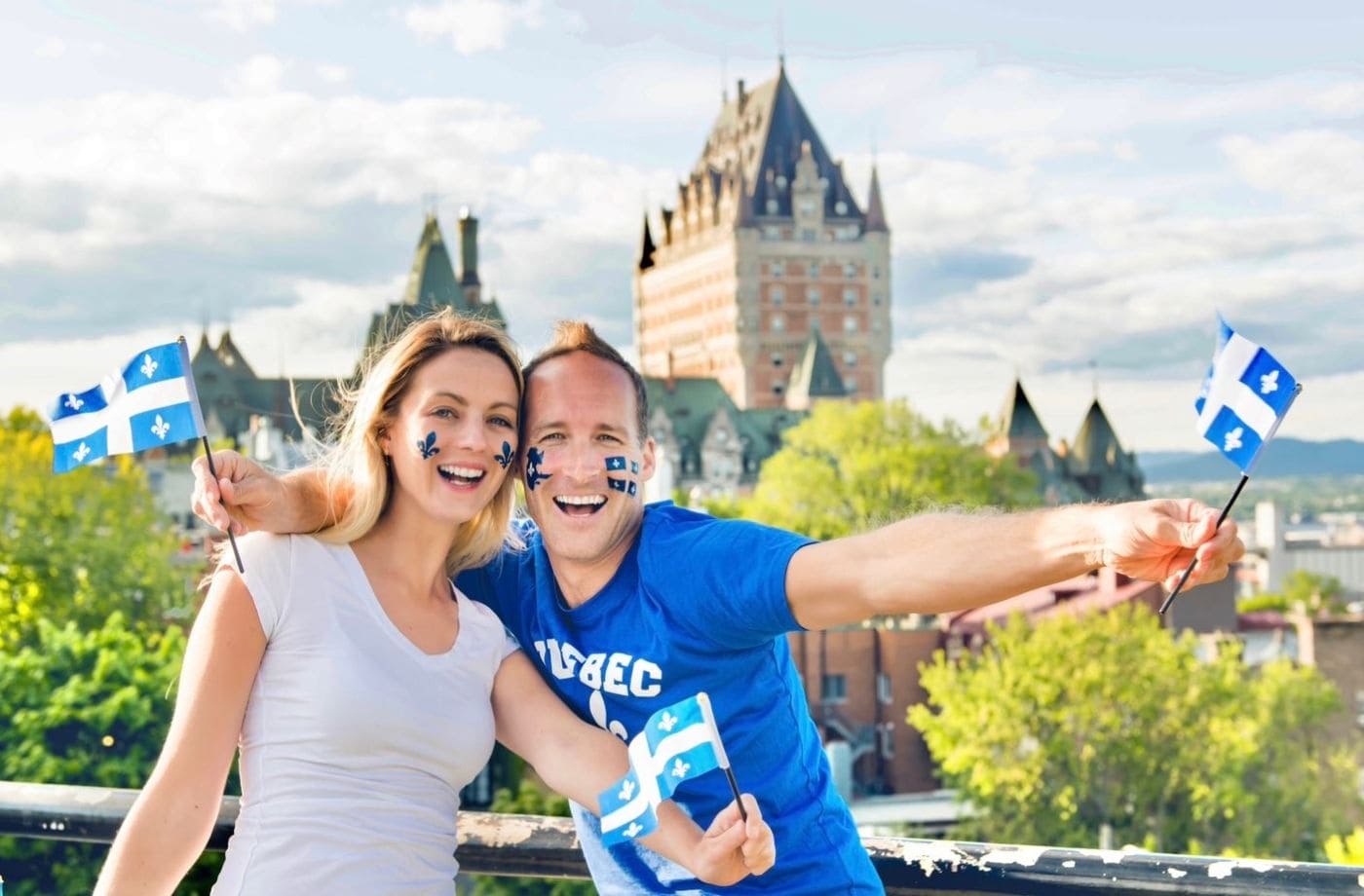 Рейтинг уровня счастья в Канаде | Наш Ванкувер Couple smiling and waving small Quebec flags on a balcony with a château-like hotel in the background; face painted in blue fleur-de-lis symbols.
