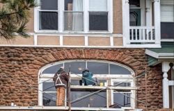 Two workers on scaffolding repairing a large arched window in a brick building.