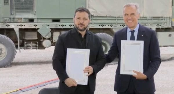Two men stand outdoors in front of a large military vehicle, each holding a framed certificate.