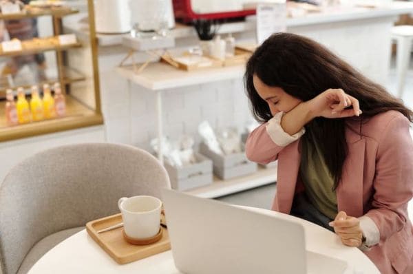 Woman in a pink blazer sits at a cafe table with a laptop, leaning her head on her arm.
