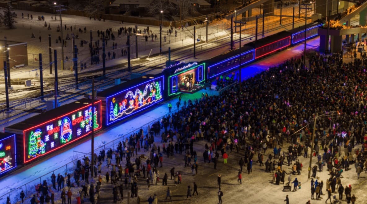 Aerial night view of a crowded street parade with a neon-lit train float and holiday panels along the rails.