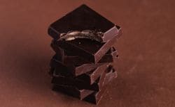 Stack of glossy dark chocolate squares stacked in a tower on a brown background, with a bite taken from the top piece