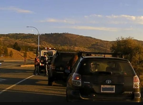 Black Toyota SUV parked on a highway with its rear hatch open, a person nearby, and a tow truck in the background against brown hills under a blue sky.