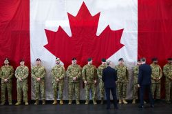 Line of Canadian soldiers in camouflage uniforms standing at attention in front of a large Canadian flag backdrop; two officials in suits face them.