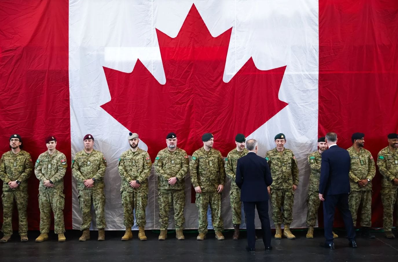 США назвали главной угрозой суверенитету Канады в Арктике | Наш Ванкувер Line of Canadian soldiers in camouflage uniforms standing at attention in front of a large Canadian flag backdrop; two officials in suits face them.