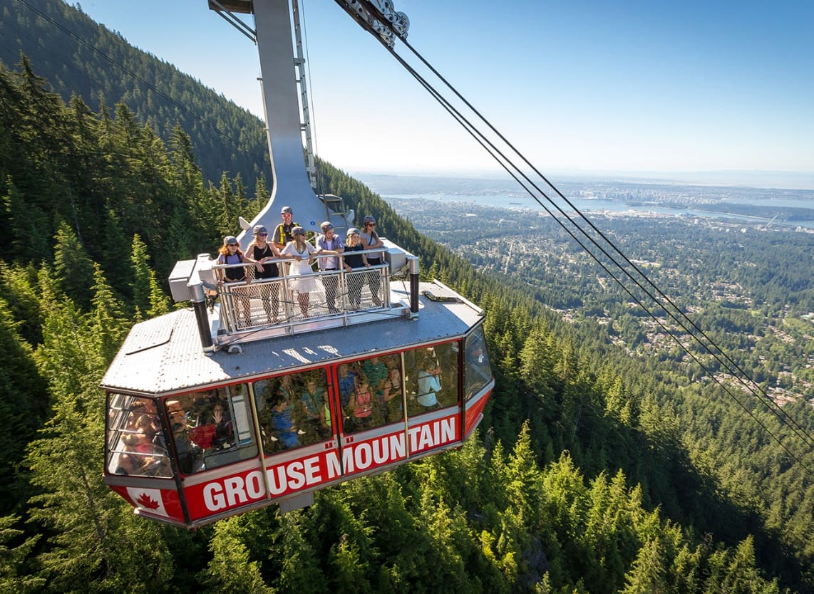 старые кабины grouse | Наш Ванкувер Cable car gondola labeled 'GROUSE MOUNTAIN' carrying passengers over a forested mountain with a city skyline in the distance.