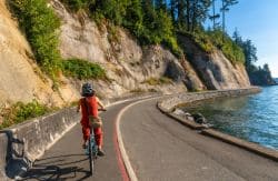 Cyclist wearing a red outfit and helmet rides along a curved coastal road beside a rocky cliff and blue water to the right.