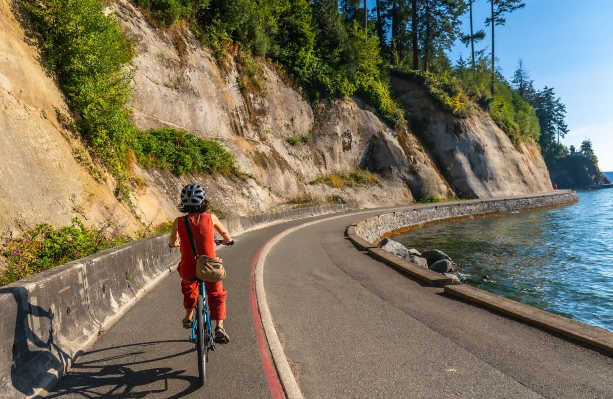 Cyclist wearing a red outfit and helmet rides along a curved coastal road beside a rocky cliff and blue water to the right.