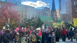 Large crowd of protesters in a city square, many holding red signs and flags, with a church and tall buildings visible in the background.