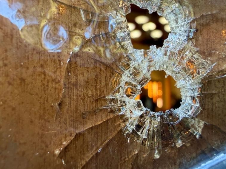 Close-up of a shattered glass surface with two bullet-hole-like holes and radiating cracks, revealing a warm-lit interior beyond.