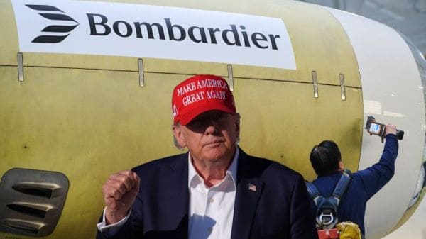 Man in a dark suit and white shirt wearing a red MAGA hat stands in front of a Bombardier aircraft; a person with a camera films in the background.