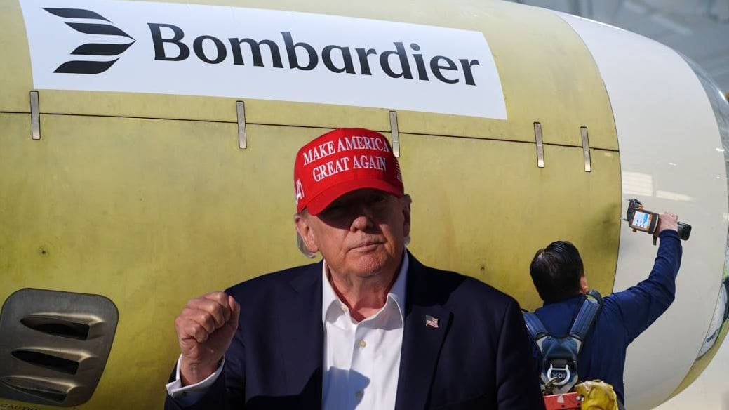 Man in a dark suit and white shirt wearing a red MAGA hat stands in front of a Bombardier aircraft; a person with a camera films in the background.
