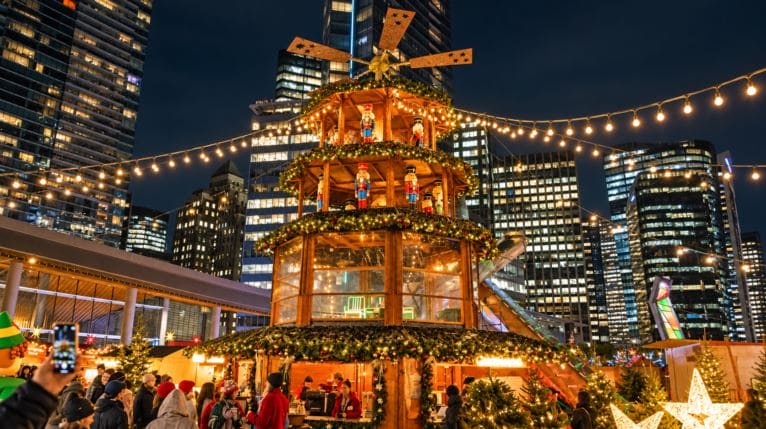 Nighttime city Christmas market with a tall wooden, multi-tiered lit Christmas tree centerpiece, string lights, and shoppers in the square.