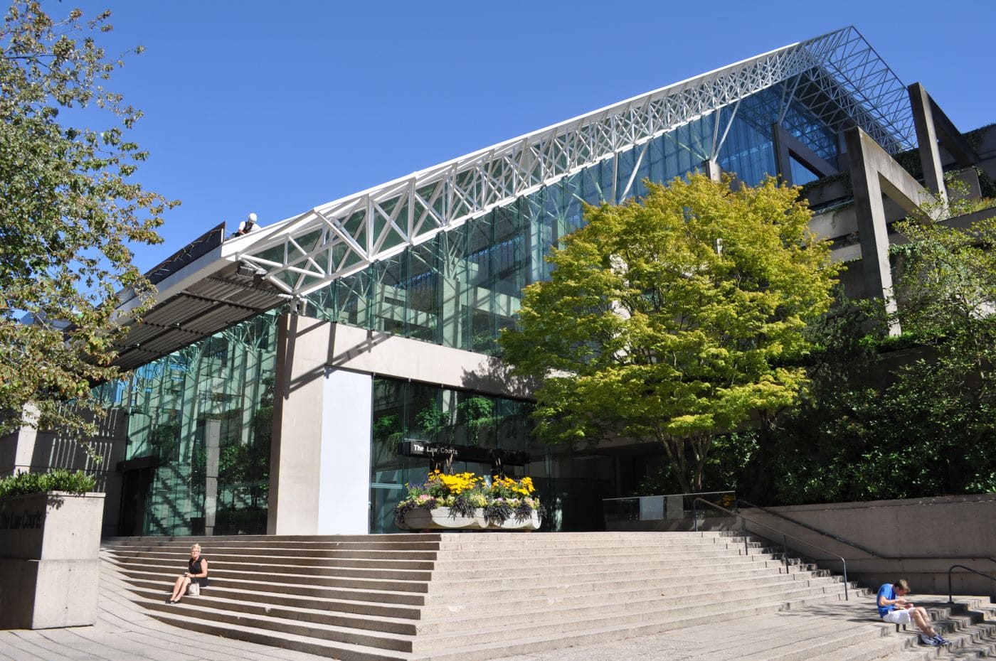 Vancouver_-_law_courts_01 | Наш Ванкувер Modern glass building with a white steel lattice canopy and wide concrete steps, flanked by trees and flowers; a person sits on the steps.