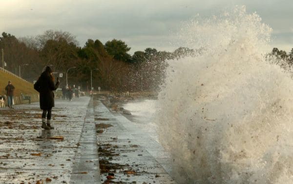 Person in a dark coat stands on a wet promenade as a large wave crashes against the seawall, spraying water into the air; others walk in the distance.