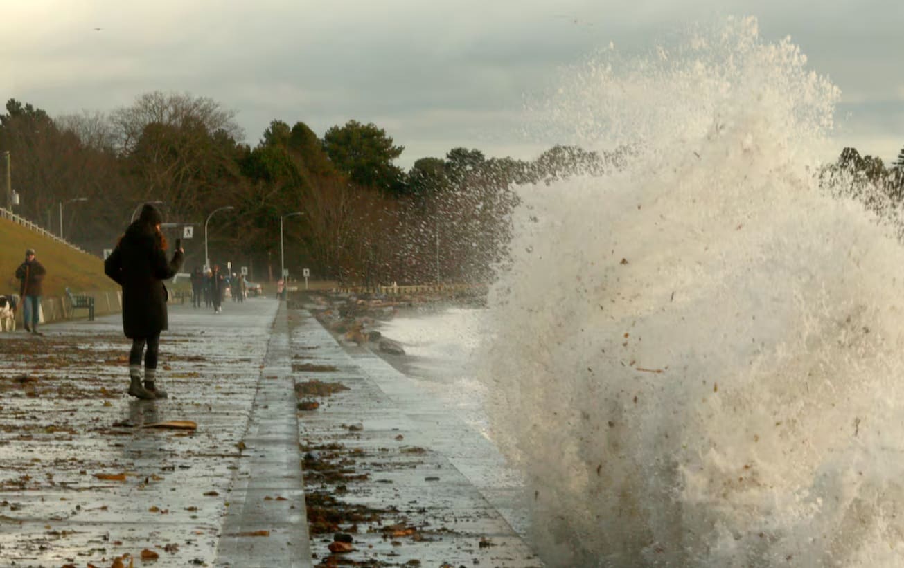волна кельвина | Наш Ванкувер Person in a dark coat stands on a wet promenade as a large wave crashes against the seawall, spraying water into the air; others walk in the distance.