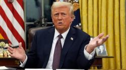 Man in a dark suit and red tie speaks at a desk with flags behind him, gesturing with open hands.
