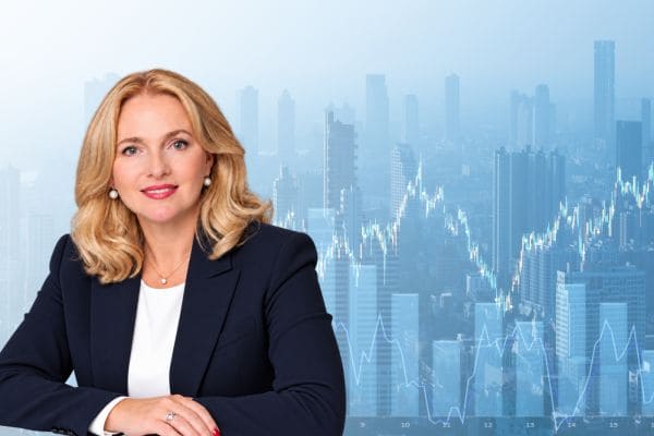 Professional blonde woman in a navy blazer at a desk, with a blue city skyline and stock charts in the background.