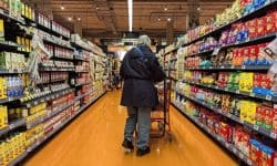 Senior shopper with a cart walks down a bright supermarket aisle lined with cereal boxes on both sides.
