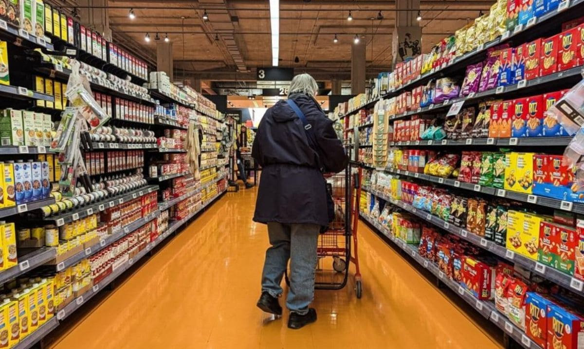 выплаты на продукты | Наш Ванкувер Senior shopper with a cart walks down a bright supermarket aisle lined with cereal boxes on both sides.
