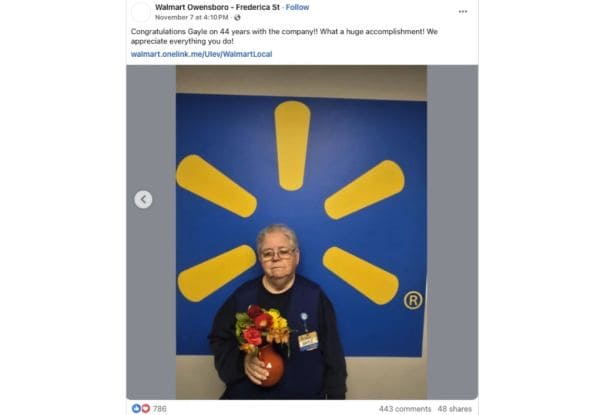 Older man with a name badge holding a bouquet of flowers in front of a blue Walmart backdrop with the yellow Walmart sunburst logo, celebrating an anniversary.