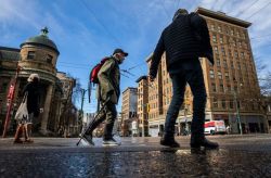 Pedestrians cross a wet city street on a sunny day; a man with a red backpack uses a walking pole while others pass by.