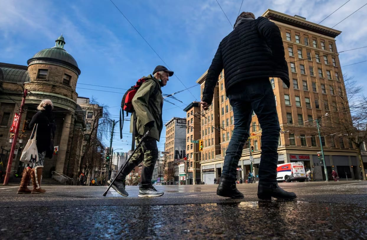Pedestrians cross a wet city street on a sunny day; a man with a red backpack uses a walking pole while others pass by.